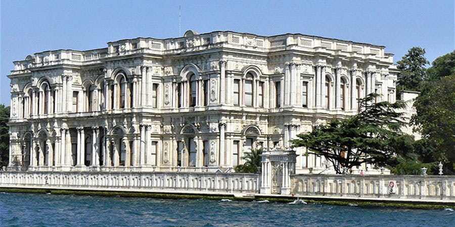 Un grande edificio bianco decorato con finestre ad arco e dettagli intricati, situato vicino a uno specchio d'acqua con un cielo azzurro e limpido.