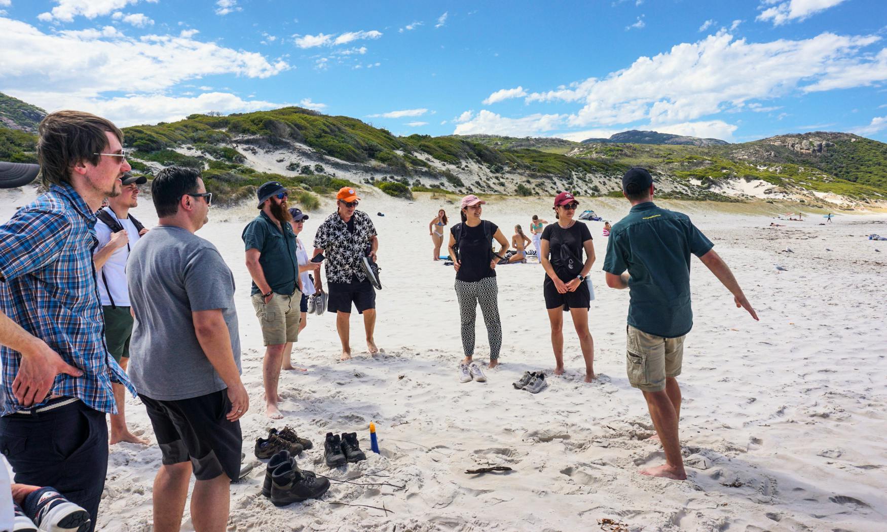 Μια ομάδα ανθρώπων που μιλάνε σε μια αμμώδη παραλία, με καταπράσινους αμμόλοφους και γαλάζιο ουρανό με διάσπαρτα σύννεφα στο παρασκήνιο.
