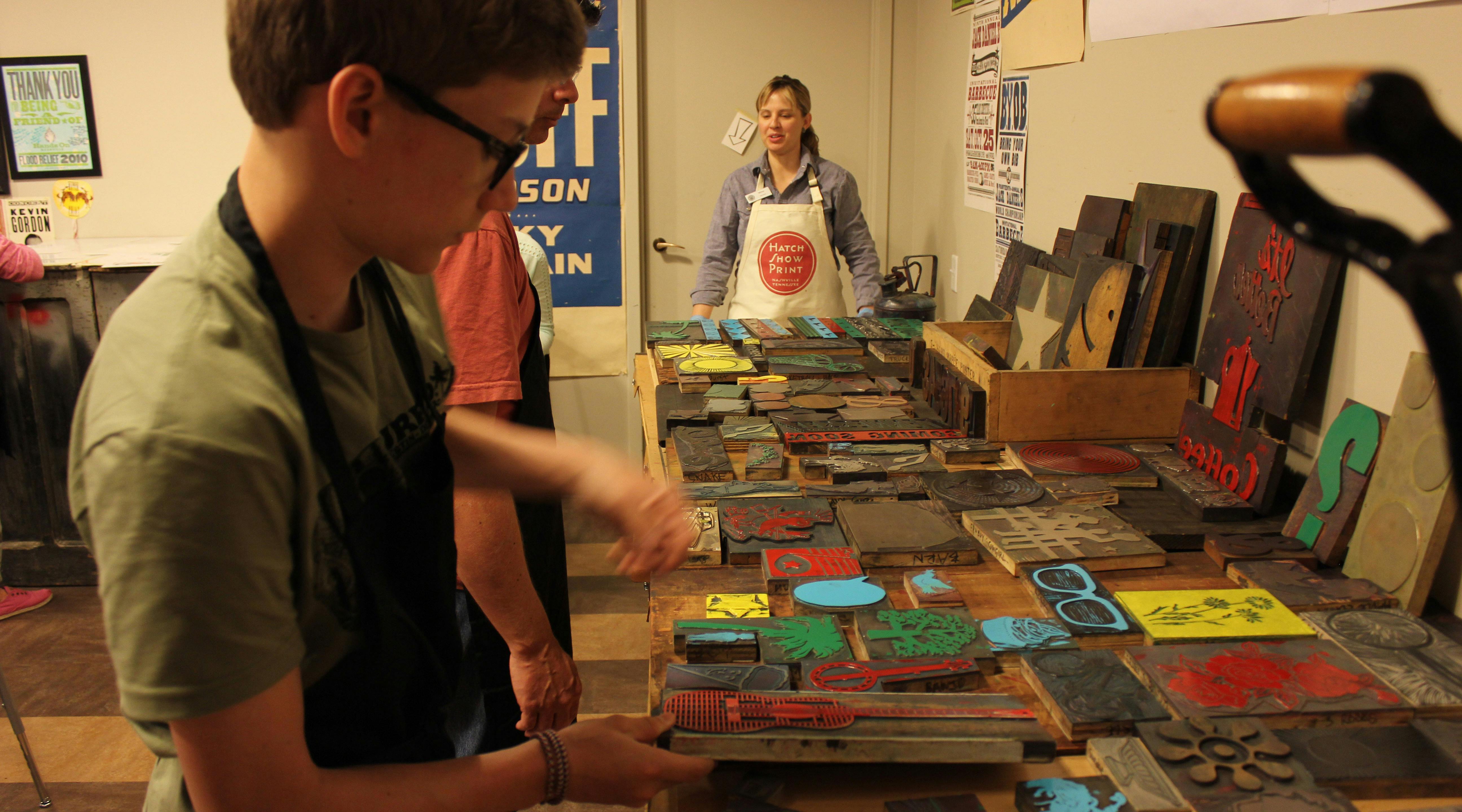 People working around a table filled with colorful printing blocks. A person wearing an apron stands in the background.