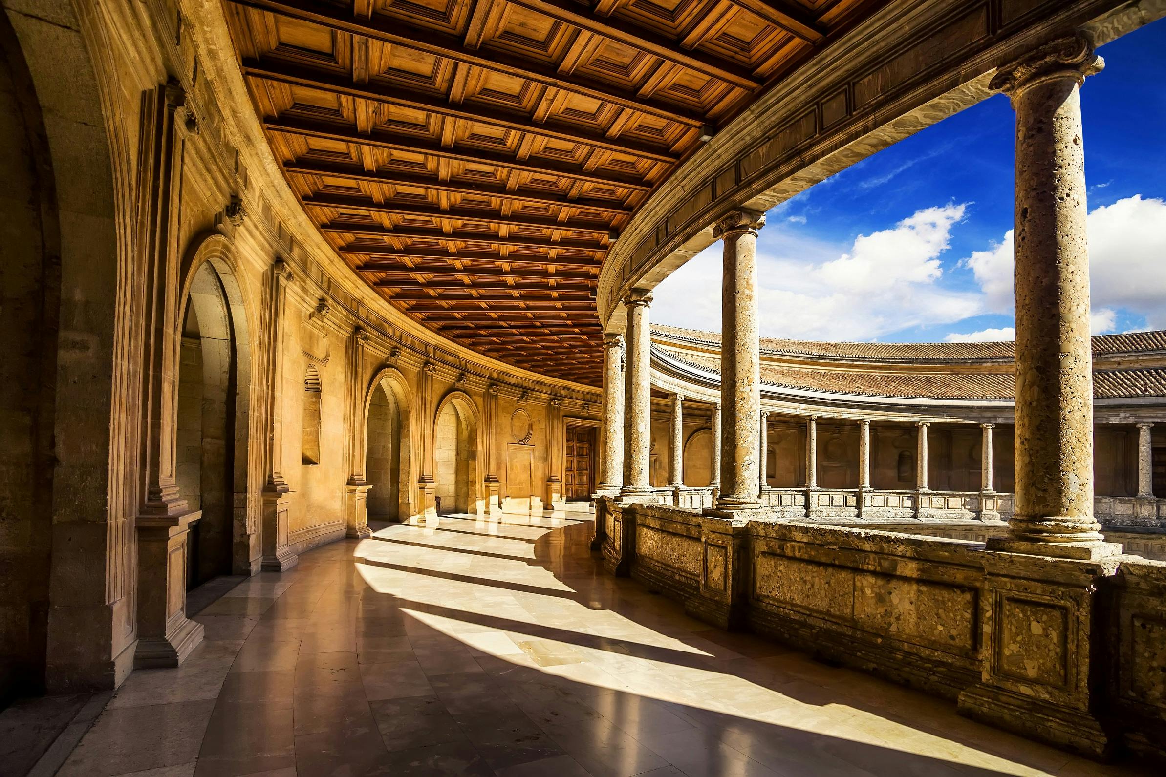 A sunlit, circular stone courtyard with arches and columns, featuring a wooden coffered ceiling and a blue sky above.