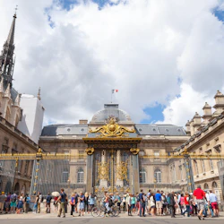 En folkmassa samlas framför en utsmyckad grind prydd med gulddekorationer, vilket leder till en historisk byggnad med en fransk flagga.