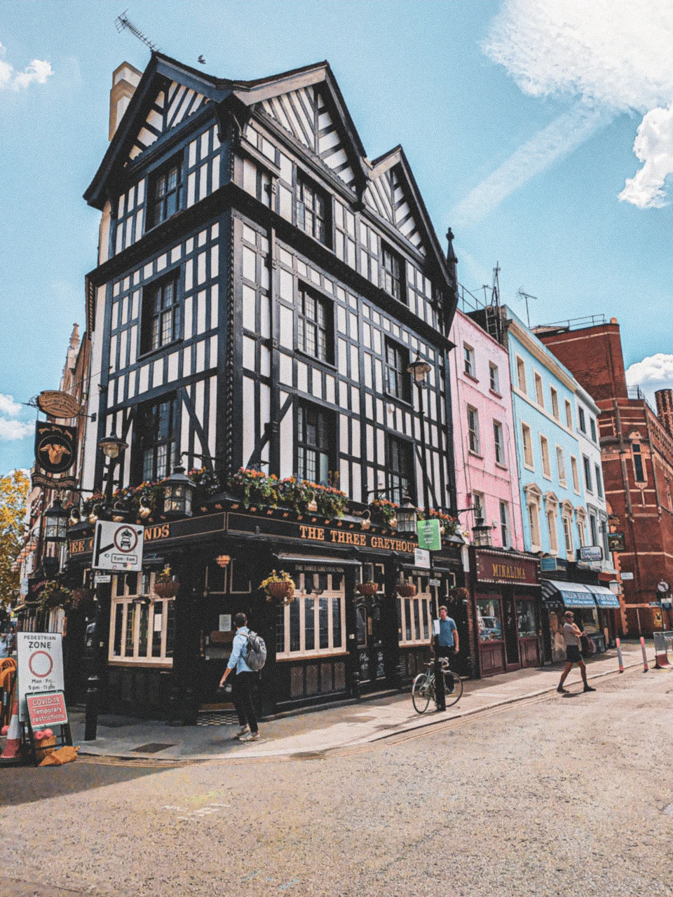 A corner pub named "The Three Greyhounds" with half-timbered architecture, adjacent colorful buildings, and pedestrians on the street.