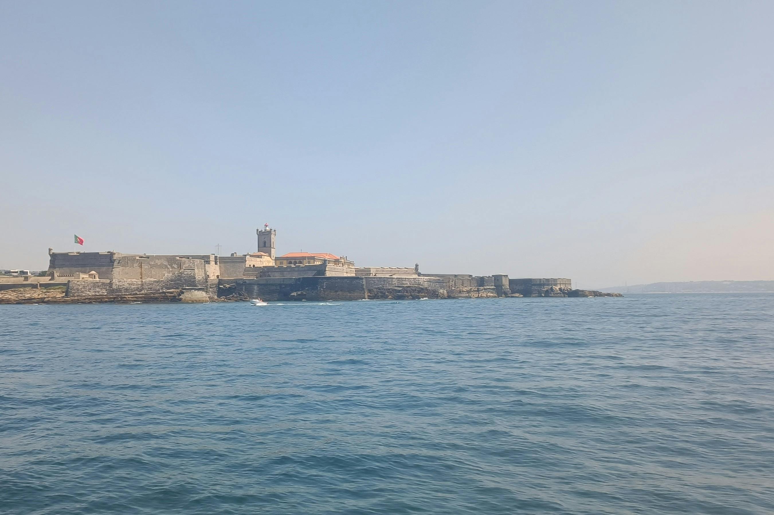 Ένα πέτρινο φρούριο με έναν πύργο σε μια ακτογραμμή που φαίνεται από τη θάλασσα κάτω από έναν καταγάλανο ουρανό.