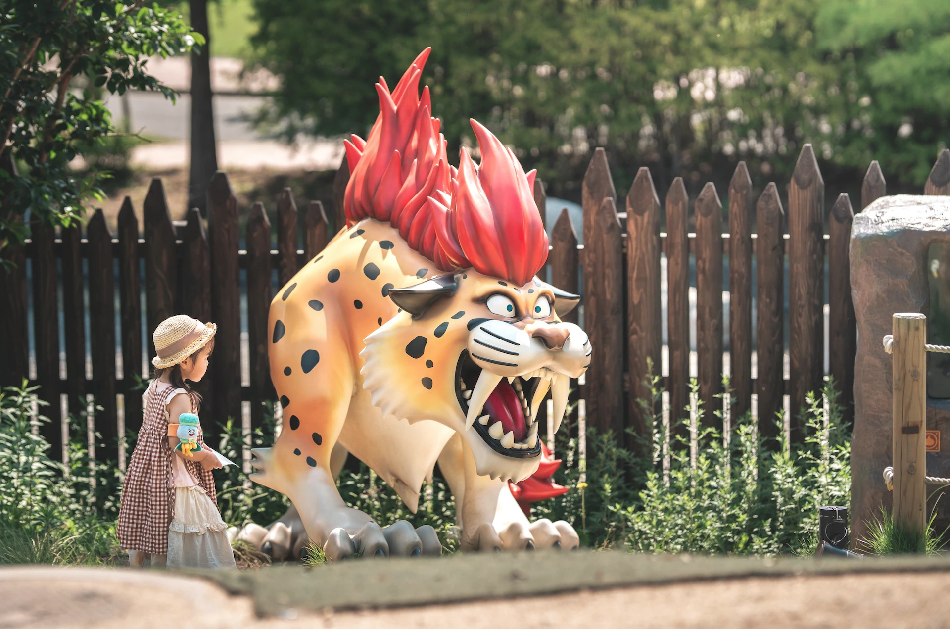 A child stands near a large statue of a roaring, red-maned, spotted feline in front of a wooden fence in an outdoor setting.