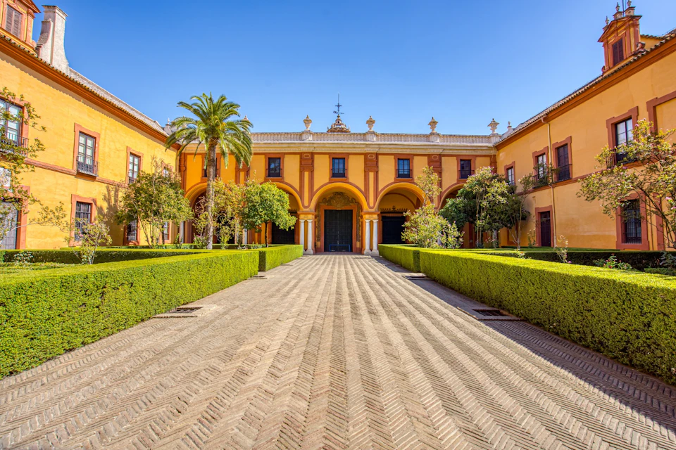 A symmetrical courtyard with yellow walls, arches, hedges, and trees under a clear blue sky.