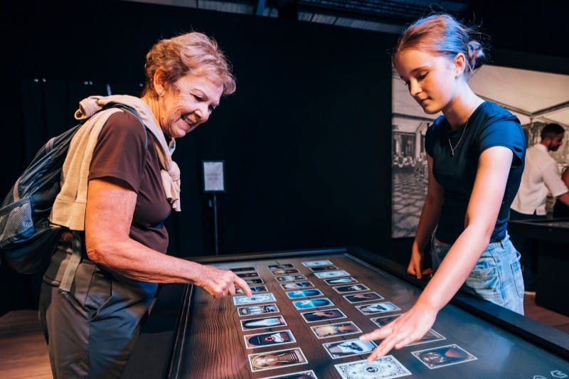 两个人在昏暗的房间里与显示卡片的触摸屏进行交互。 背景中有另一个人。