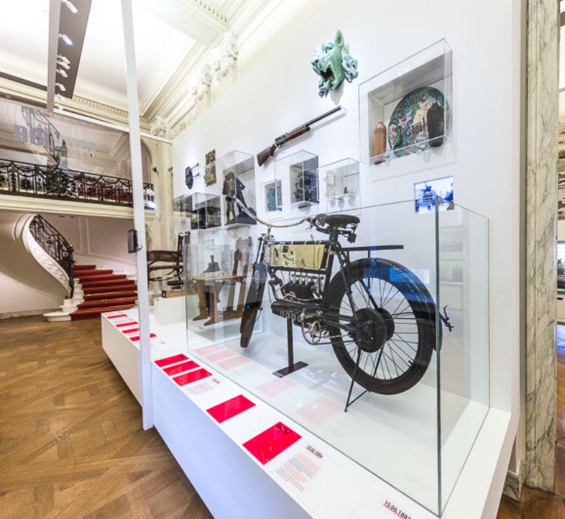 Exposició del museu amb una motocicleta vintage en una caixa de vidre, objectes històrics a la paret i una escala de fons.