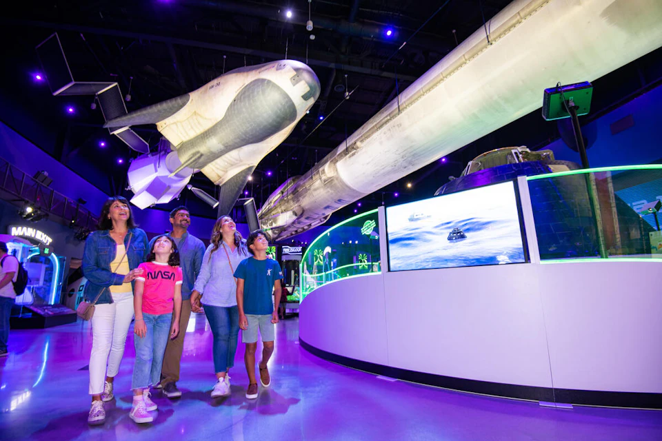 A family gazes up at a suspended space shuttle in a museum exhibit, surrounded by interactive displays and vivid lighting.