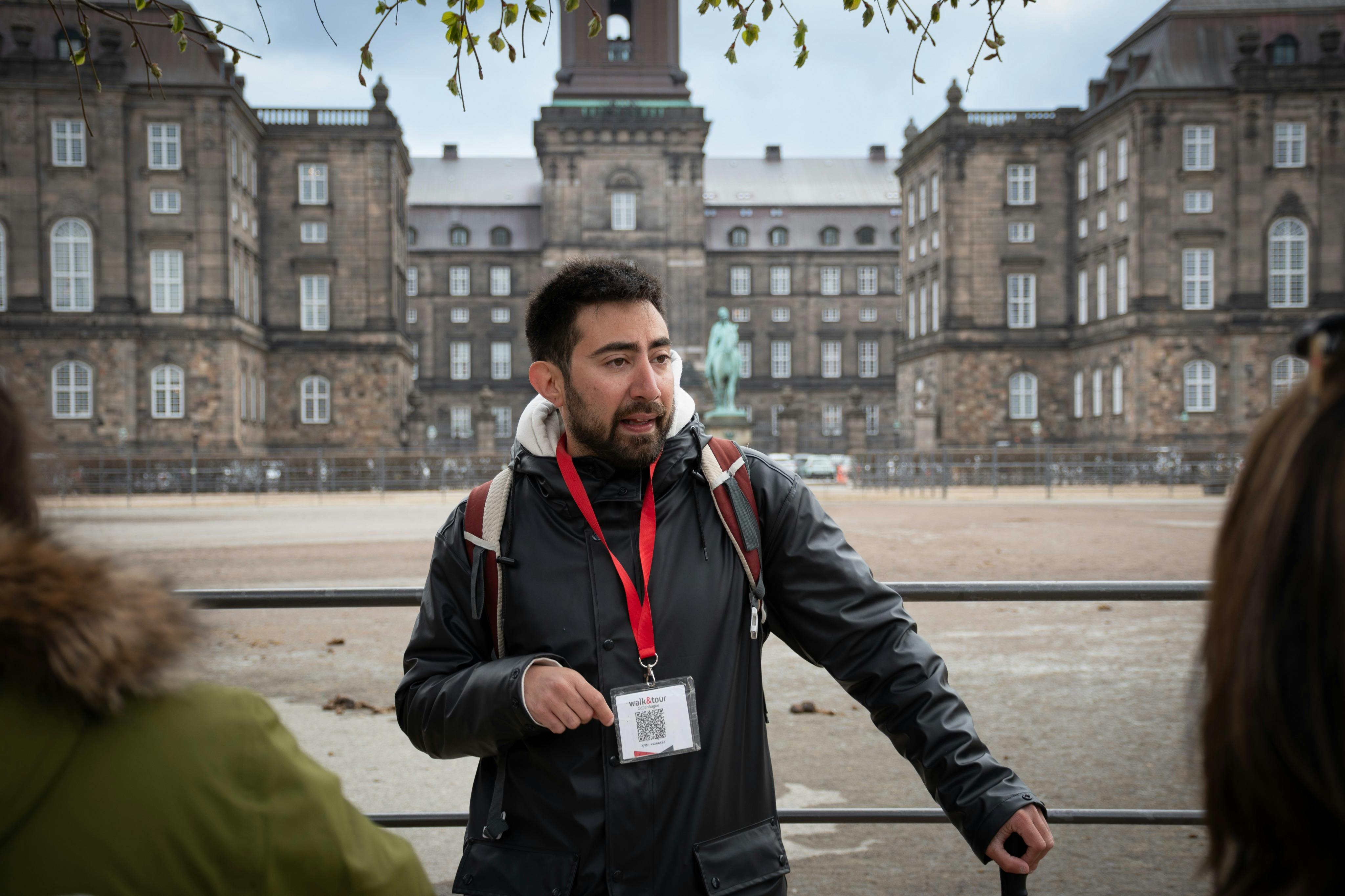 一位戴着挂绳的男士在一座历史悠久的大型建筑前说话；另一个人在前景中部分可见。