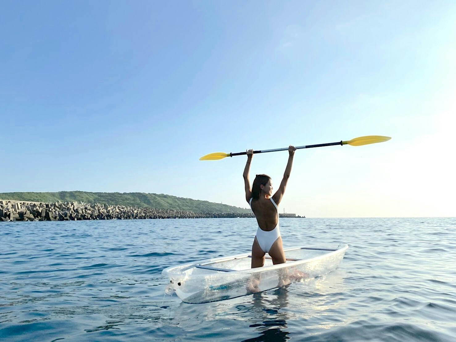 A person in a white swimsuit holding a paddle above their head while sitting in a transparent kayak on calm blue water.