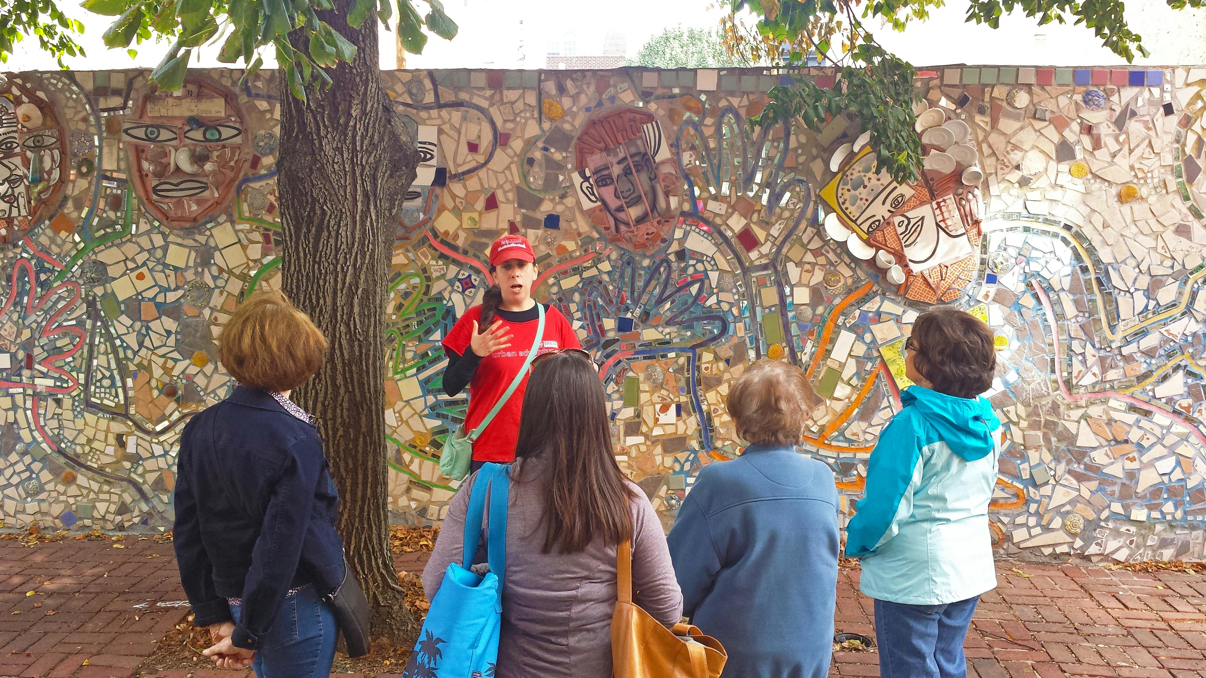 Een gids met een rode hoed en overhemd spreekt vier mensen aan voor een kleurrijke mozaïekmuur onder een boom.