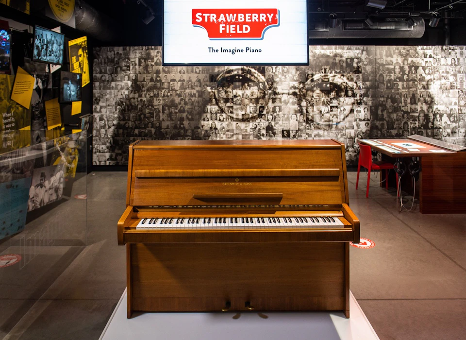 A wooden piano in a museum exhibit with a sign above reading "Strawberry Field: The Imagine Piano" and a monochrome photo backdrop.