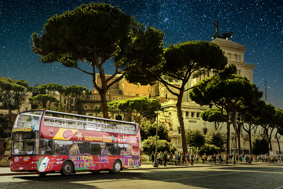 A red double-decker tour bus drives past a historic Roman building with tall trees and a starry night sky above.