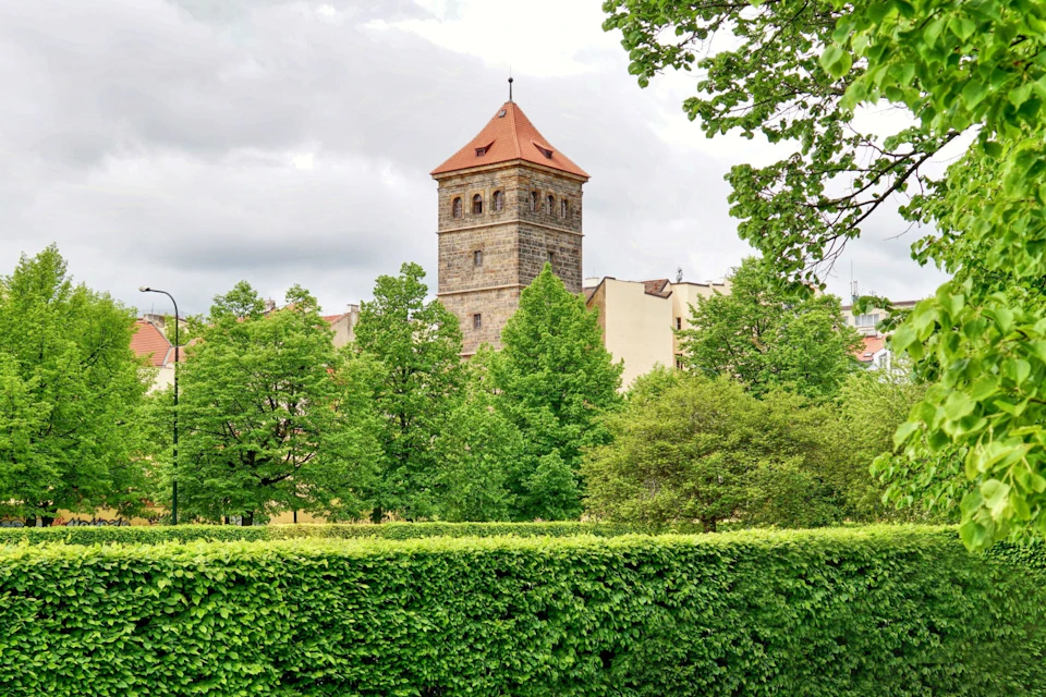 A stone tower with a red roof stands among lush green trees and bushes under a cloudy sky.