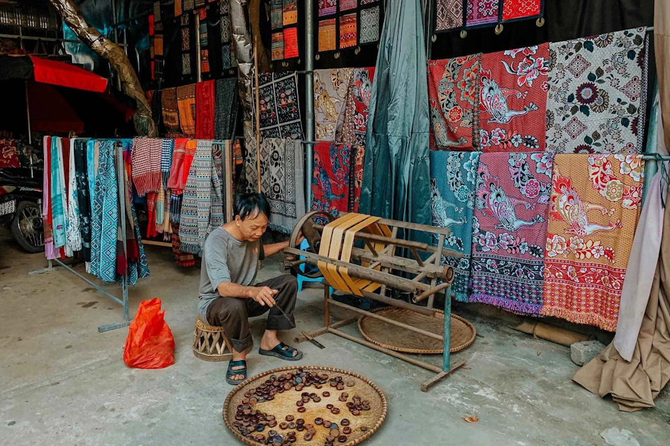 An older man sits on a small stool next to a basket of chestnuts in front of a colorful textile stall.