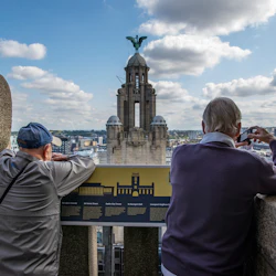 Två män har utsikt över en stadsbild från en hög utsiktspunkt, med en man som tar ett foto och en hög byggnad med en staty synlig i bakgrunden.