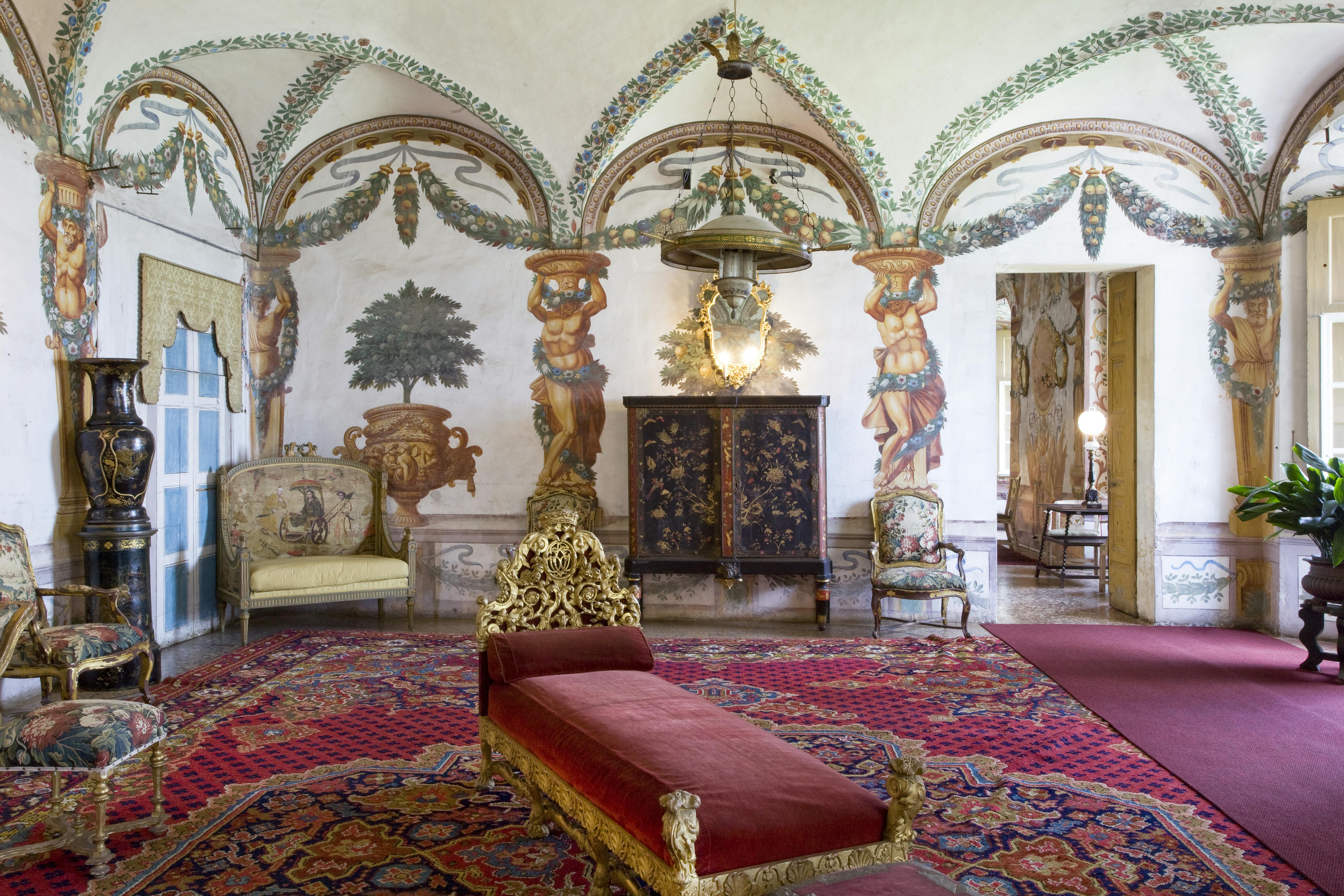 Ornate room with a red chaise longue, tapestry chairs, a decorative cabinet, and frescoes of figures and plants on arched walls.
