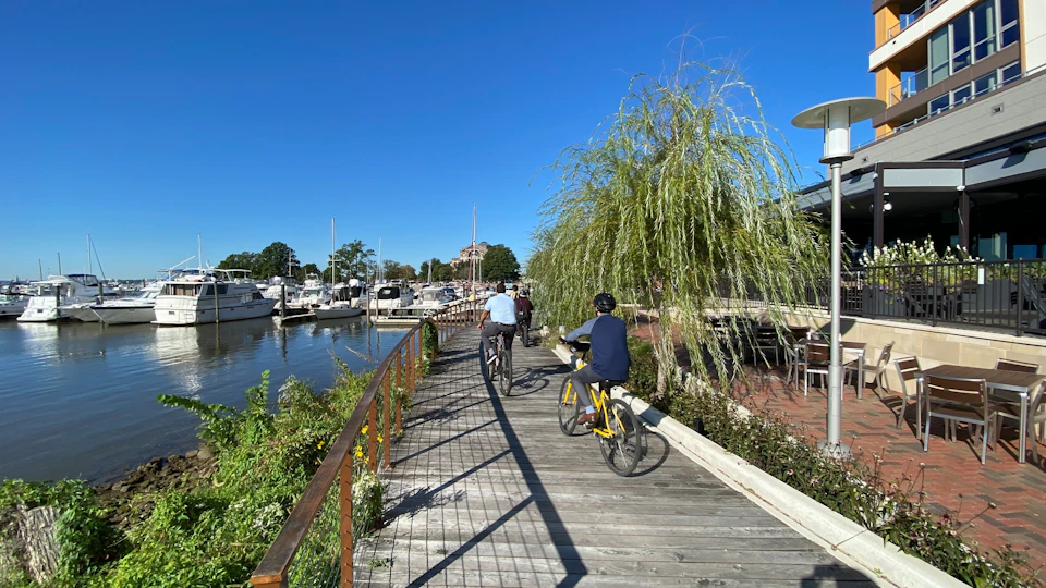 Two people bike on a wooden boardwalk beside a marina with boats docked. A willow tree and clear blue sky are in the background.