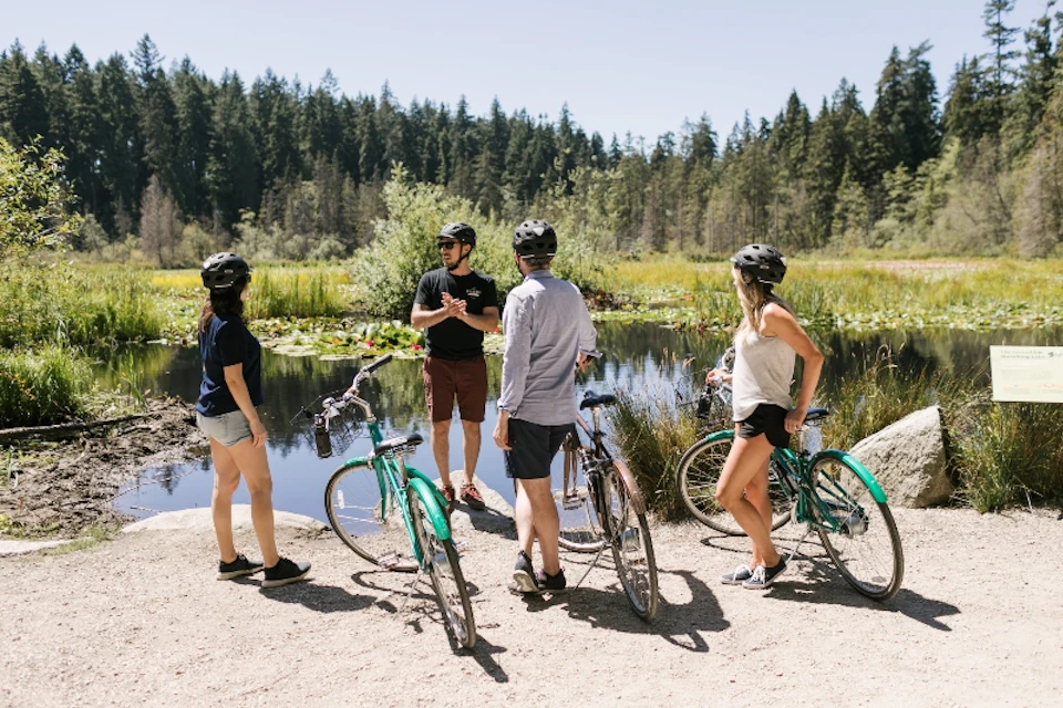 Four people with bicycles stand near a pond in a forested area, wearing helmets and casual clothing, having a conversation.