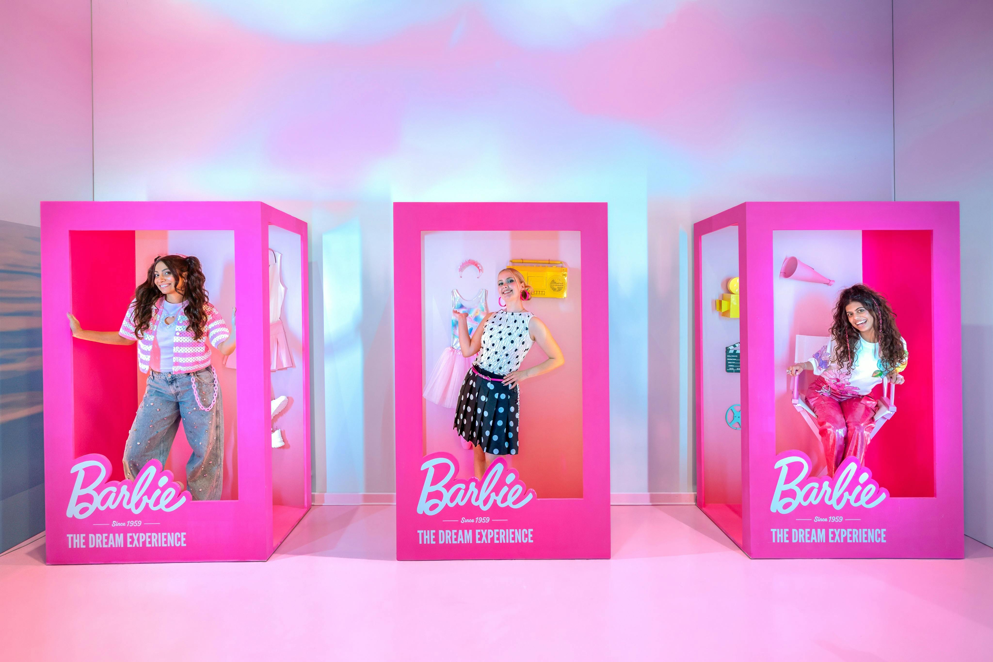 Dvě ženy pózují uvnitř růžových vitrín s motivem Barbie s nápisem „The Dream Experience“ s různými doplňky kolem sebe.
