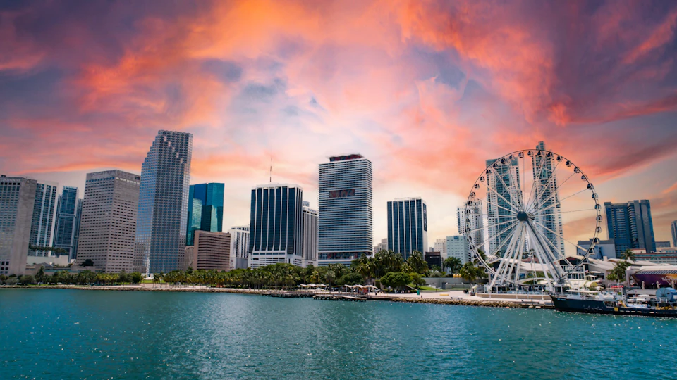 City skyline with tall buildings, a Ferris wheel, and waterfront, under a vibrant orange and pink sunset sky.