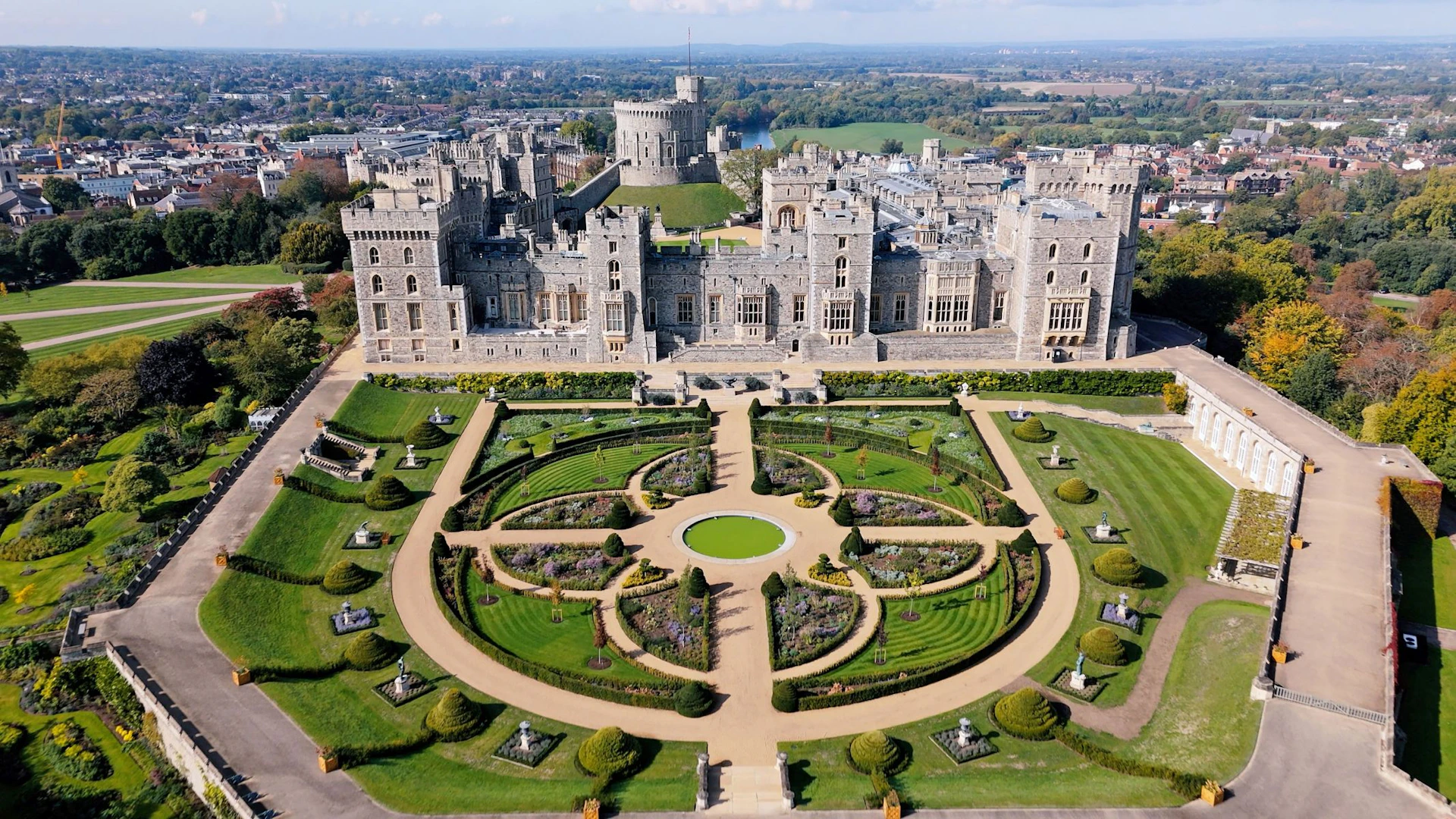 Aerial view of a large castle with intricate gardens featuring symmetrical pathways and greenery in the foreground.