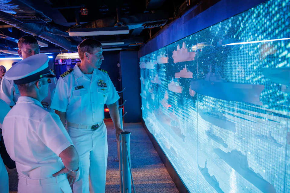 A person in a naval uniform observes a digital display wall with lit models of ships in a dark, futuristic room.