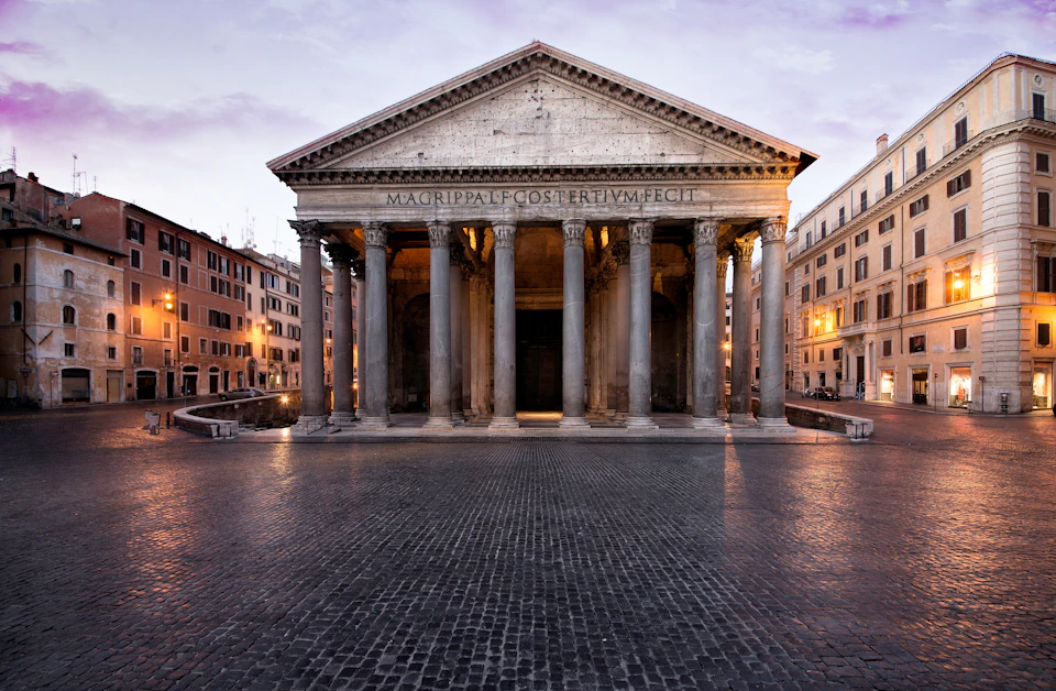 Ancient temple with large columns and inscription on the facade, surrounded by cobblestone plaza and buildings at dusk.