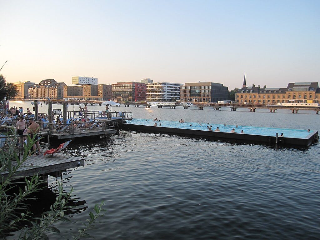 水辺の木製のデッキでリラックスしている人もいれば、水に浮かぶプールで泳いでいる人もいれば、夕暮れ時の背景にある建物もあります。