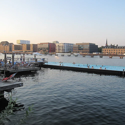 Människor som kopplar av på ett trädäck vid vattnet, med andra som simmar i en flytande pool, och byggnader i bakgrunden vid solnedgången.