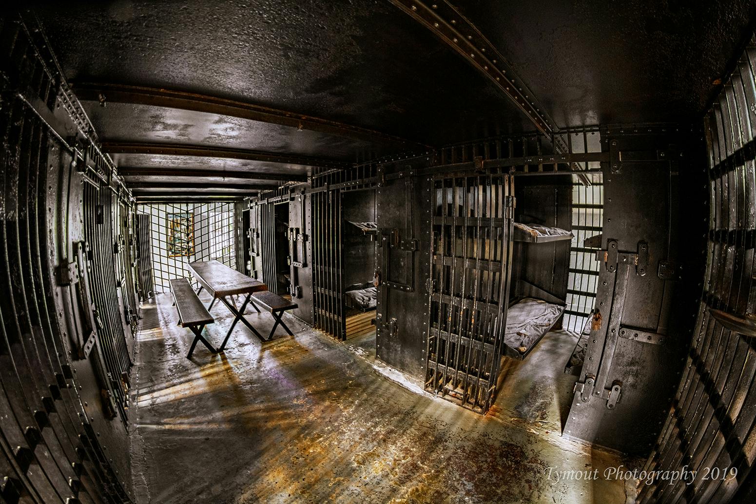 A dimly lit prison cell block with metal bars, bunk beds, and a central wooden bench and table.