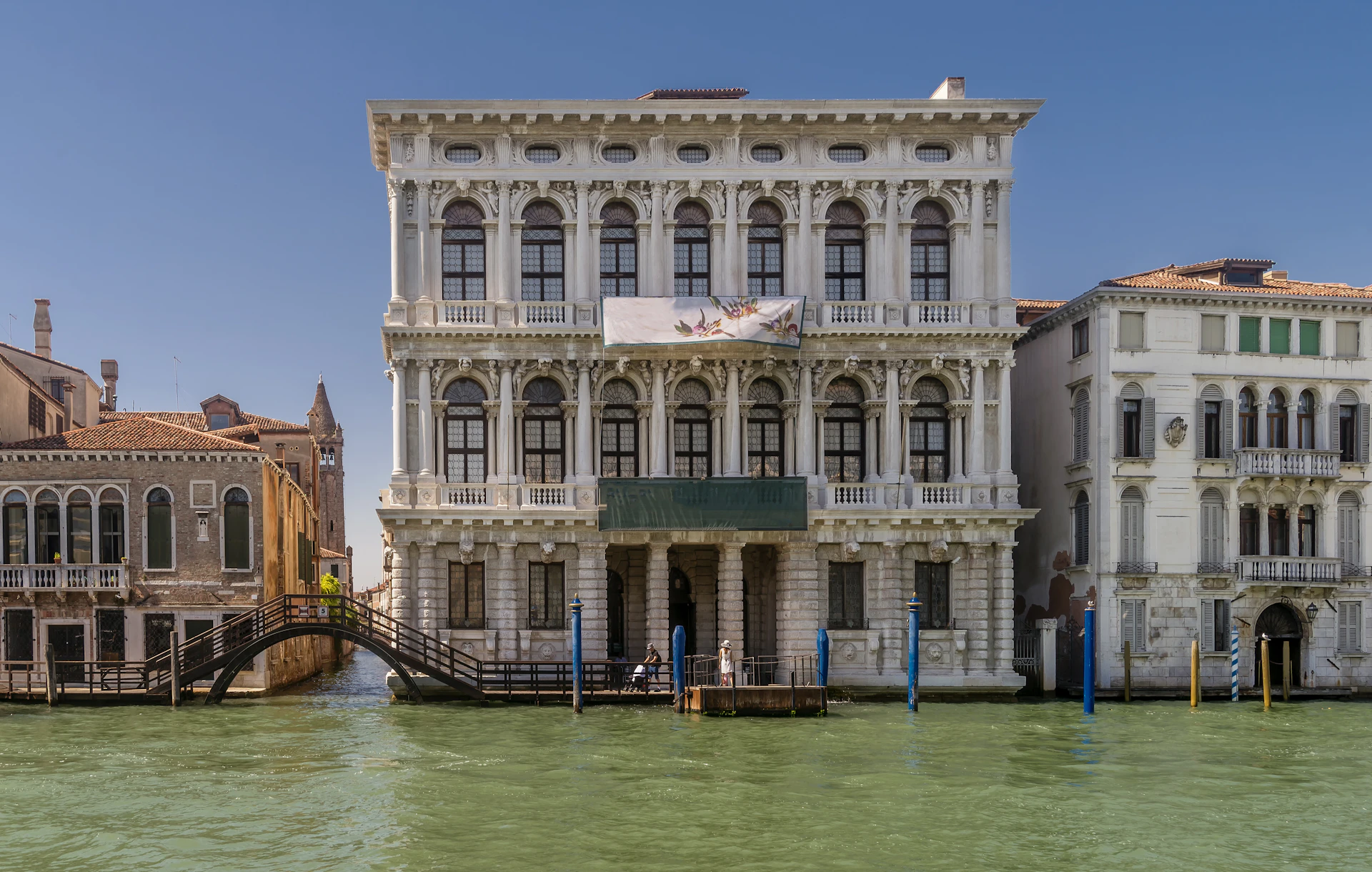 Three-story ornate white building by a canal, with arched windows, balconies, and a small wooden dock. Clear blue sky.