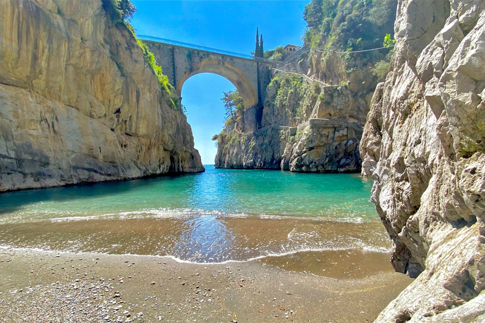 A scenic coastal view with a stone arch bridge spanning between rocky cliffs over turquoise water, near a sandy beach.