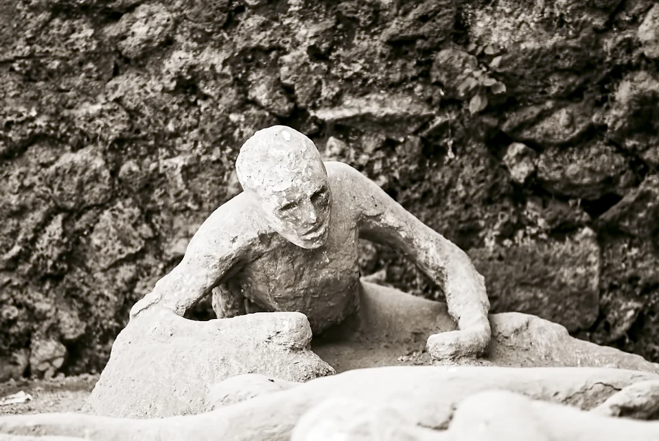 A weathered stone sculpture depicts a human figure climbing over another. The background is a rough, textured wall.