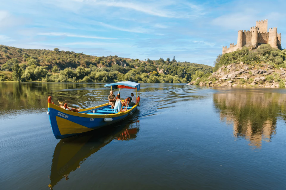 A colorful boat with a blue canopy on a calm river, surrounded by green hills and a castle visible on the right.