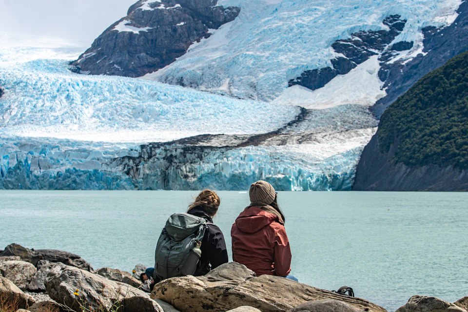 Two people sit on rocks near a turquoise lake with a large glacier and rugged mountains in the background.