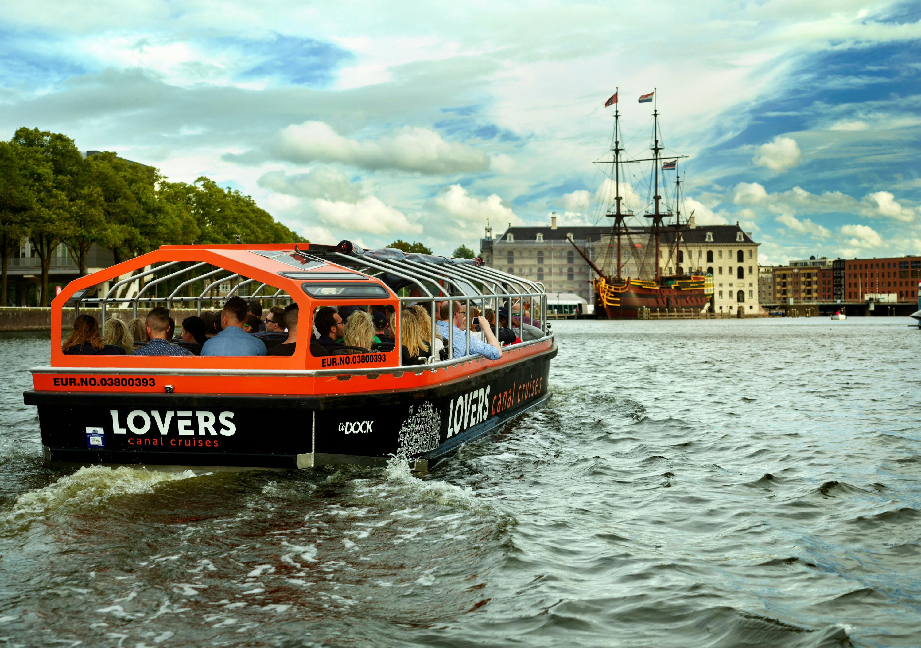 Toeristen zitten in een oranje rondvaartboot over het water, op weg naar een historisch schip en een groot gebouw onder een bewolkte lucht.