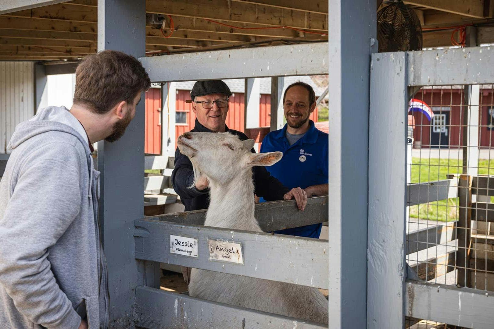 Two men petting a white goat inside a wooden pen. The pen has name tags for "Jessie" and "Angela." Red buildings in the background.