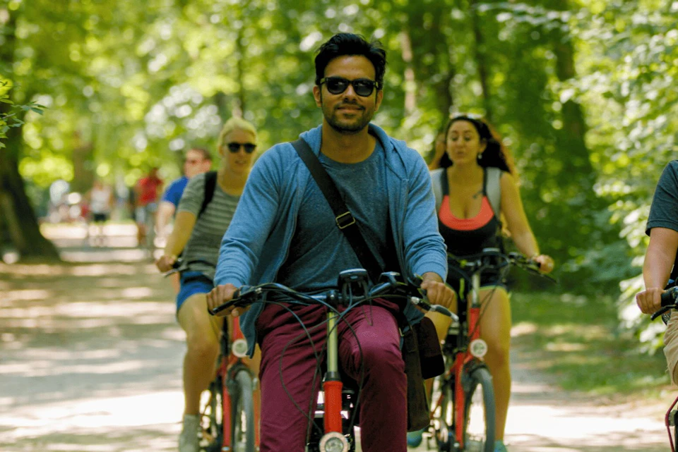 A group of people ride bicycles on a tree-lined path on a sunny day. The person in front wears sunglasses and a blue hoodie.