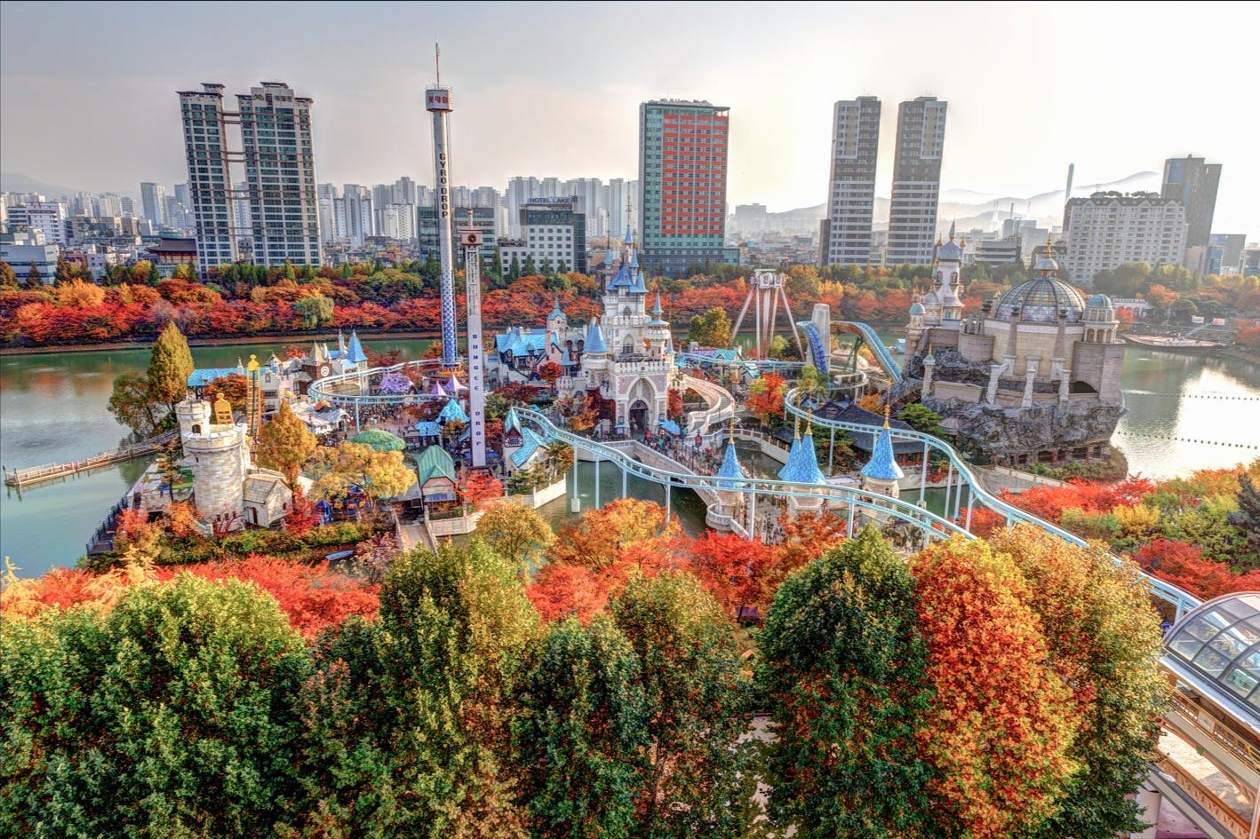 城、ジェットコースター、さまざまな乗り物を備えたテーマパークは、色とりどりの秋の木や街の建物に囲まれています。