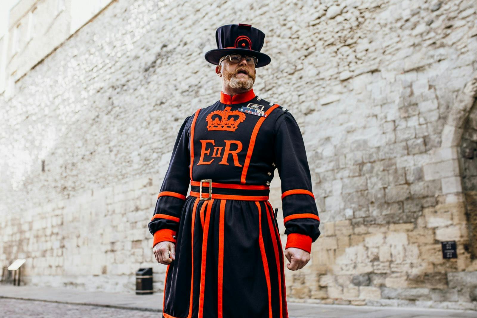 A man in traditional black and red uniform stands in front of a stone wall. His attire features a crown symbol and "ER" lettering.