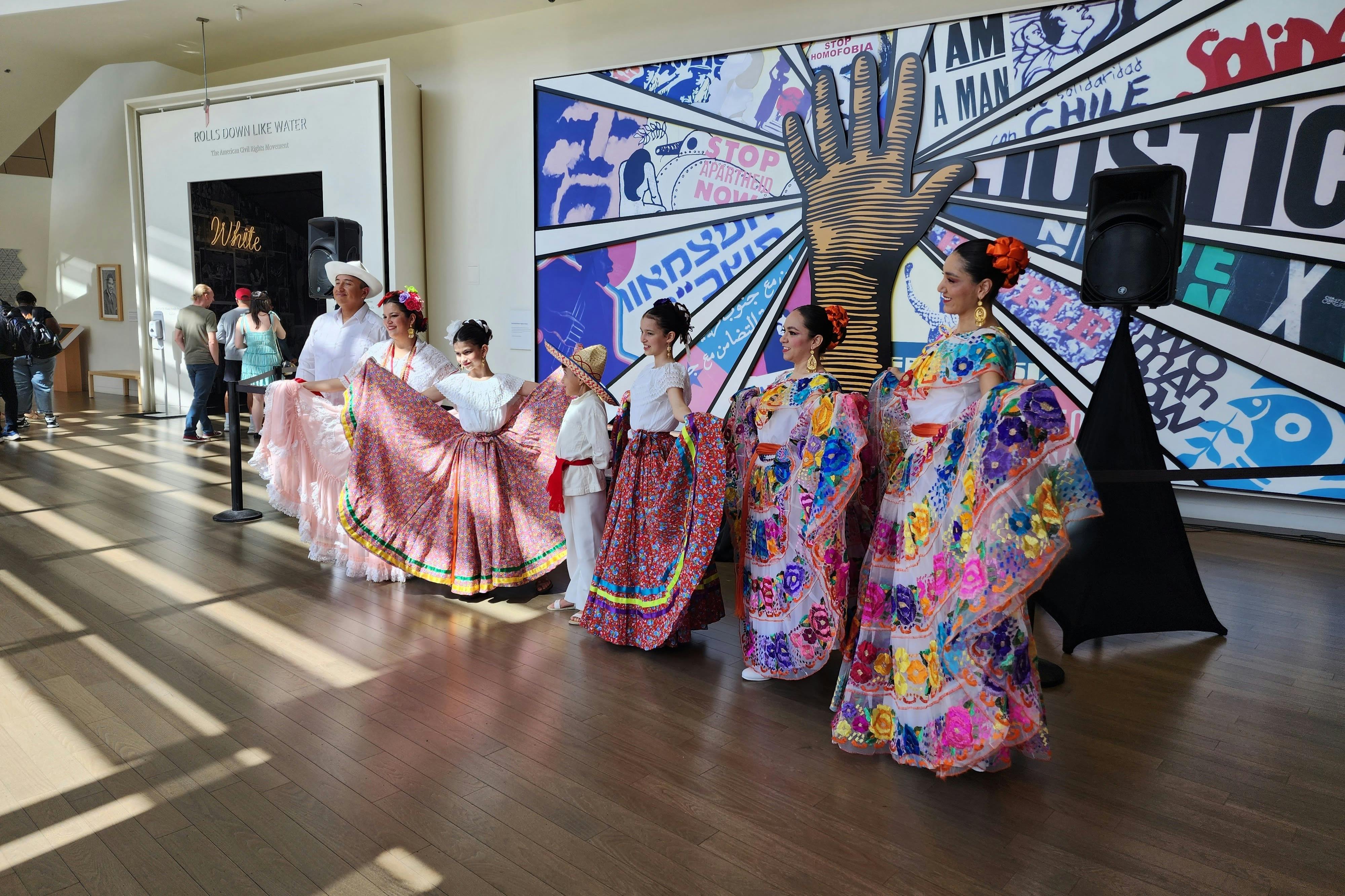 Um grupo de pessoas com trajes tradicionais coloridos actua em frente a um mural vibrante com vários textos e gráficos.