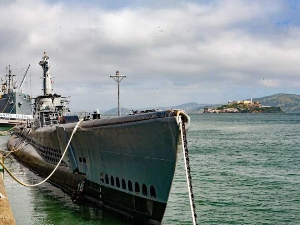 A docked submarine tied with a rope, with calm water and a distant coastline under a cloudy sky.