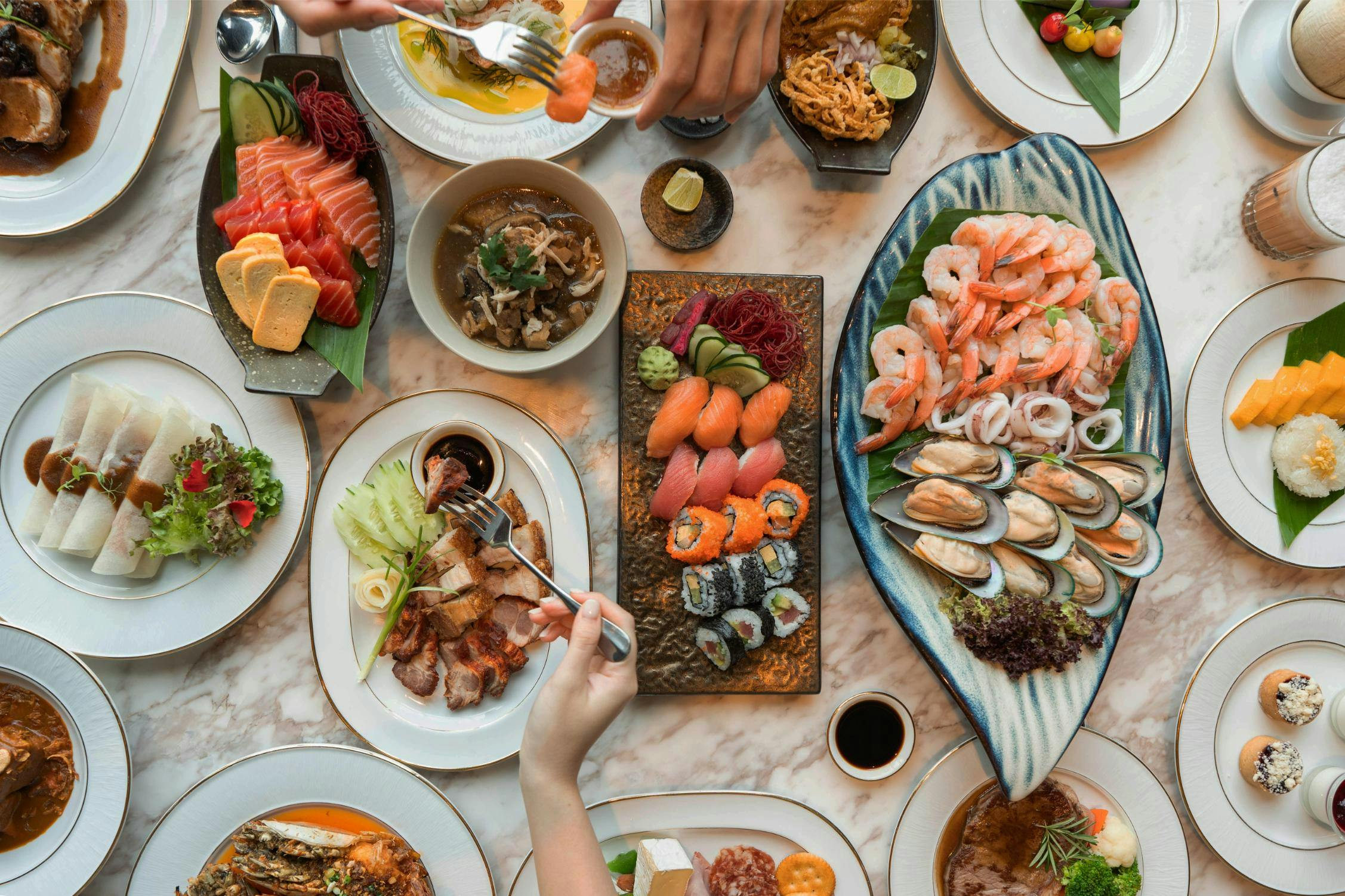 A table filled with a variety of dishes including sushi, seafood, salads, and soups, with hands reaching for food.