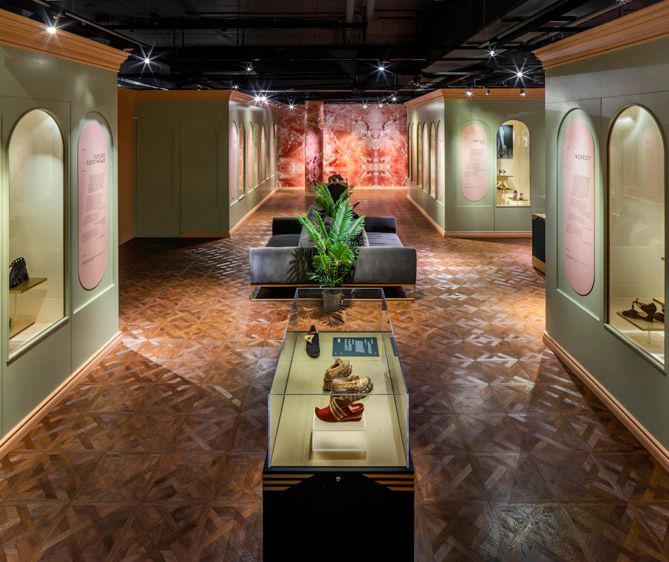 A museum exhibit with glass display cases, artifacts, informational panels, a central plant, and a patterned wood floor.