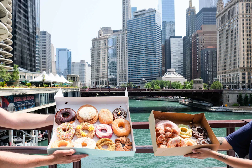 Two hands holding open boxes of assorted donuts with a city skyline and river in the background.