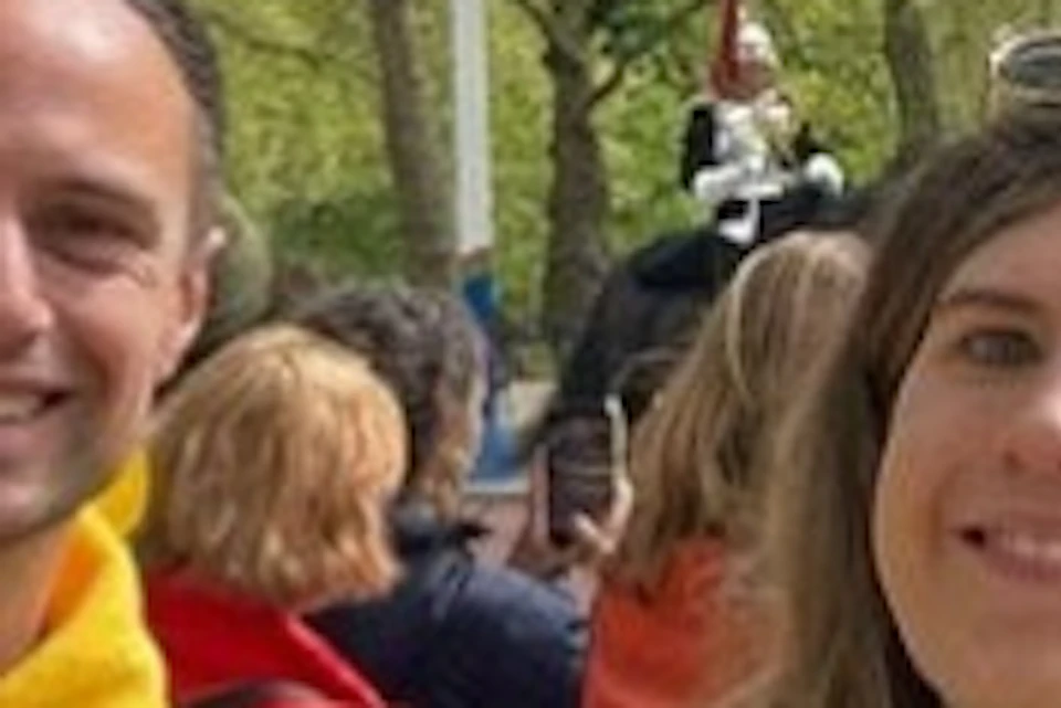 Two people holding yellow umbrellas stand outdoors in a park with trees, while a parade of horses passes in the background.