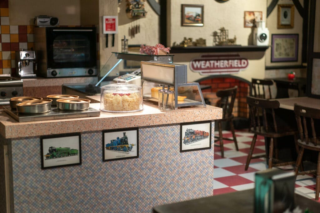 一个小咖啡馆，柜台上挂着火车的框架图片，一个蛋糕展示，墙上挂着一个“Weatherfield”标志。