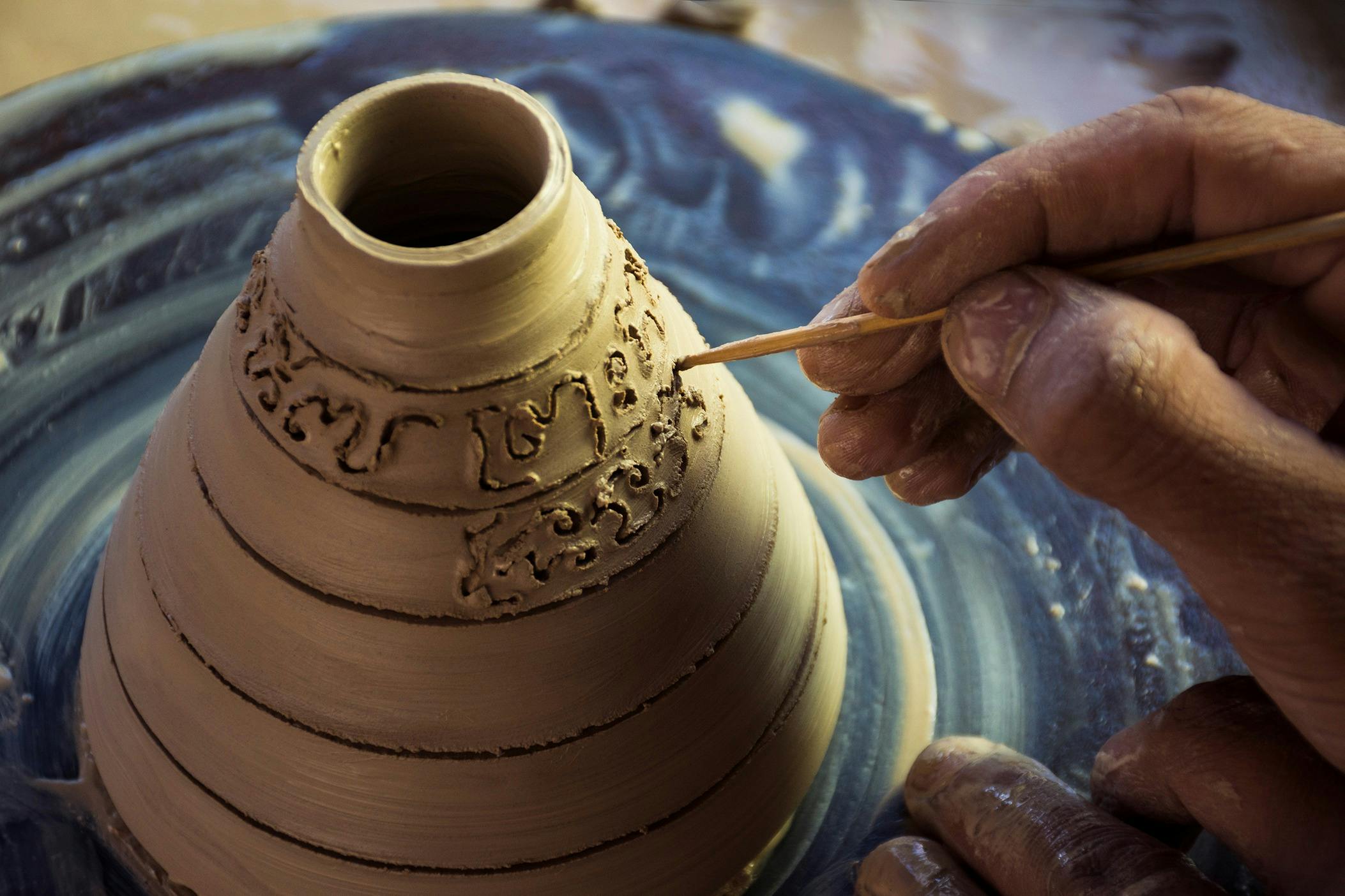 Close-up of a hand using a tool to carve designs into a clay pot on a pottery wheel.