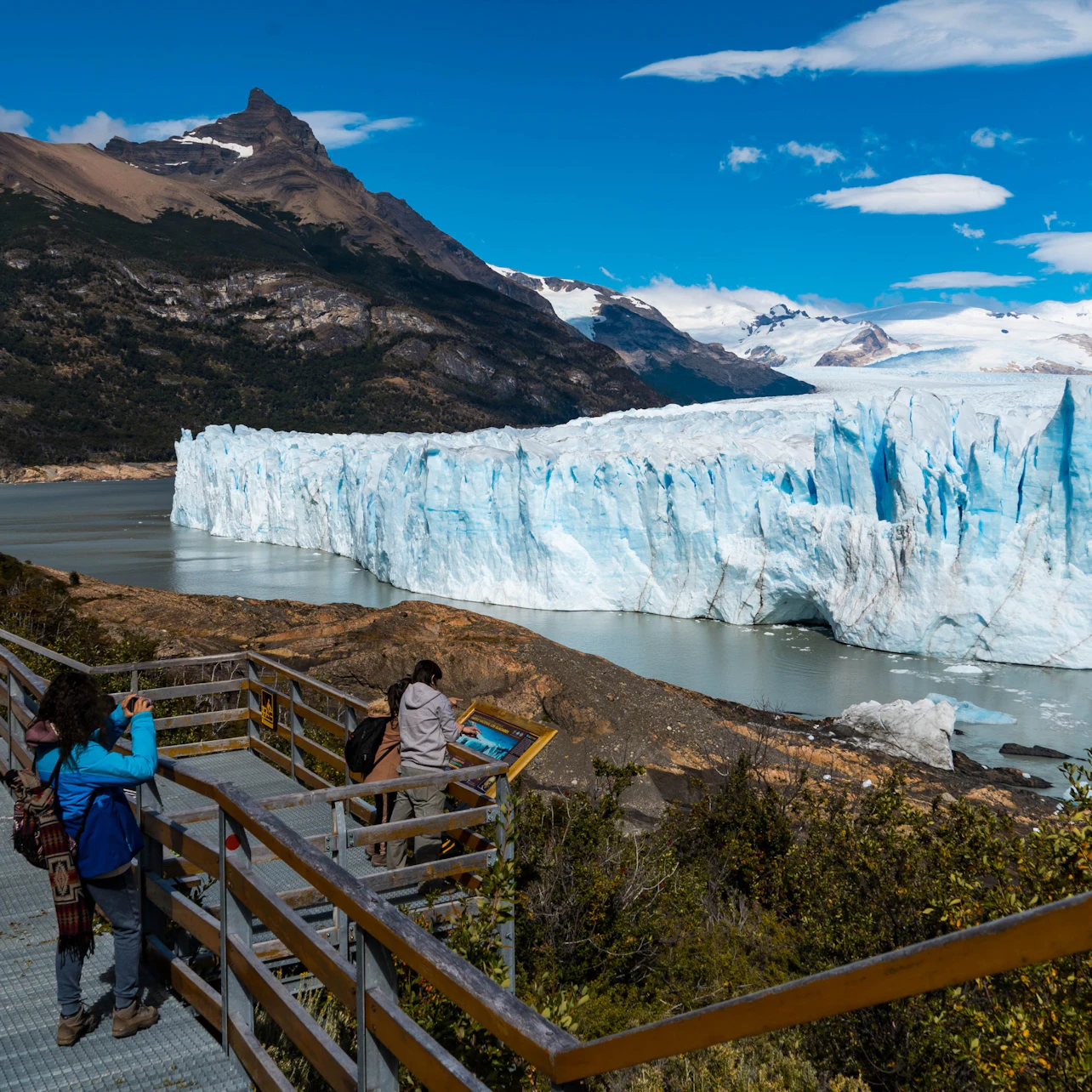 Tourists on a wooden walkway viewing a large glacier surrounded by mountains and water under a partly cloudy sky.